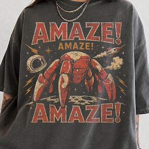 May include: A charcoal gray short-sleeve t-shirt featuring a distressed graphic print of a red robotic crab on a moon surface, a space helmet, and a rocket. The text AMAZE! is printed in large red letters above and below the central illustration.
