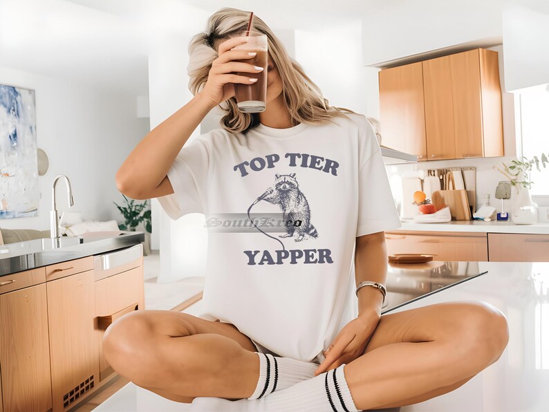 May include: White t-shirt with a graphic of a raccoon holding a microphone and the text "Top Tier Yapper".