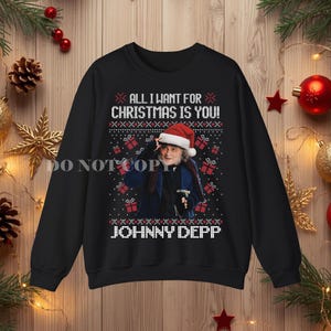 Op de afbeelding: Een zwart sweatshirt met lange mouwen en een afbeelding van Johnny Depp met een kerstmuts op. De tekst luidt ALL I WANT FOR CHRISTMAS IS YOU en JOHNNY DEPP. Het ontwerp bevat een patroon van kleine witte sneeuwvlokken en rode cadeautjes.