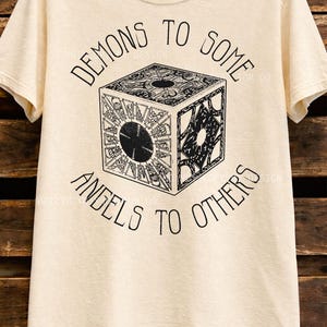 May include: Ivory-colored t-shirt with the text "DEMONS TO SOME, ANGELS TO OTHERS" in black, surrounding a detailed black and white cube graphic. The shirt is hanging on a wooden background.