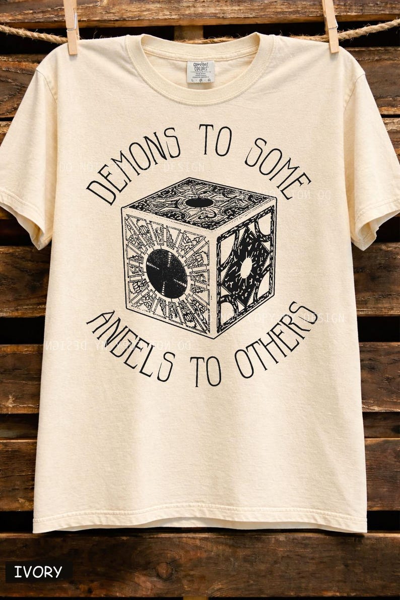 May include: Ivory-colored t-shirt with the text "DEMONS TO SOME, ANGELS TO OTHERS" in black, surrounding a detailed black and white cube graphic. The shirt is hanging on a wooden background.