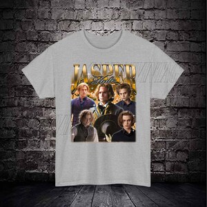 Op de afbeelding: Een grijs T-shirt met een vintage-stijl afbeelding van Jasper Hale uit de Twilight-saga. De afbeelding is een collage van afbeeldingen van Jasper Hale, met de tekst "Jasper Hale" in gouden letters.