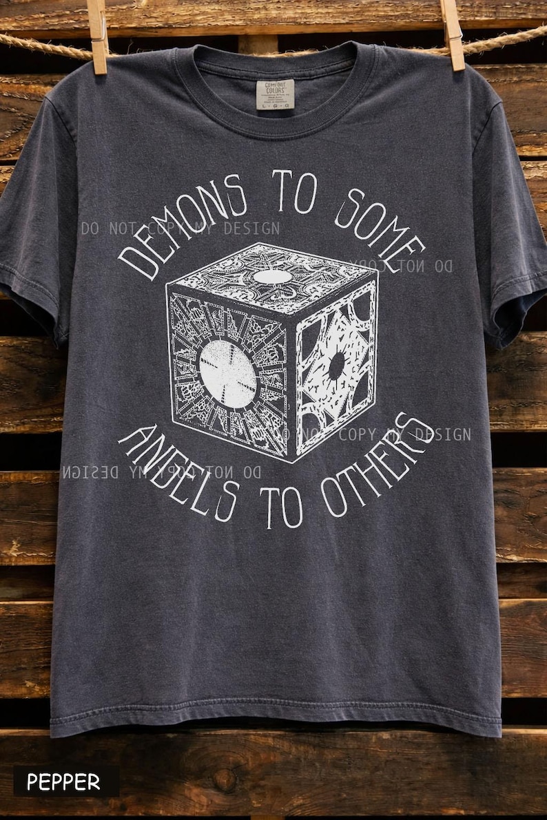 May include: Dark gray t-shirt with the text "DEMONS TO SOME, ANGELS TO OTHERS" in a curved font around a detailed, white graphic of a puzzle box. The shirt is hanging on a wooden background.