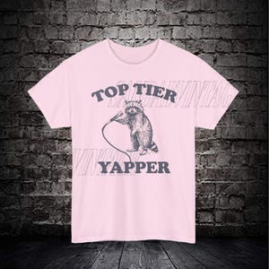 May include: Pink t-shirt with a graphic of a raccoon holding a microphone and the text "Top Tier Yapper".