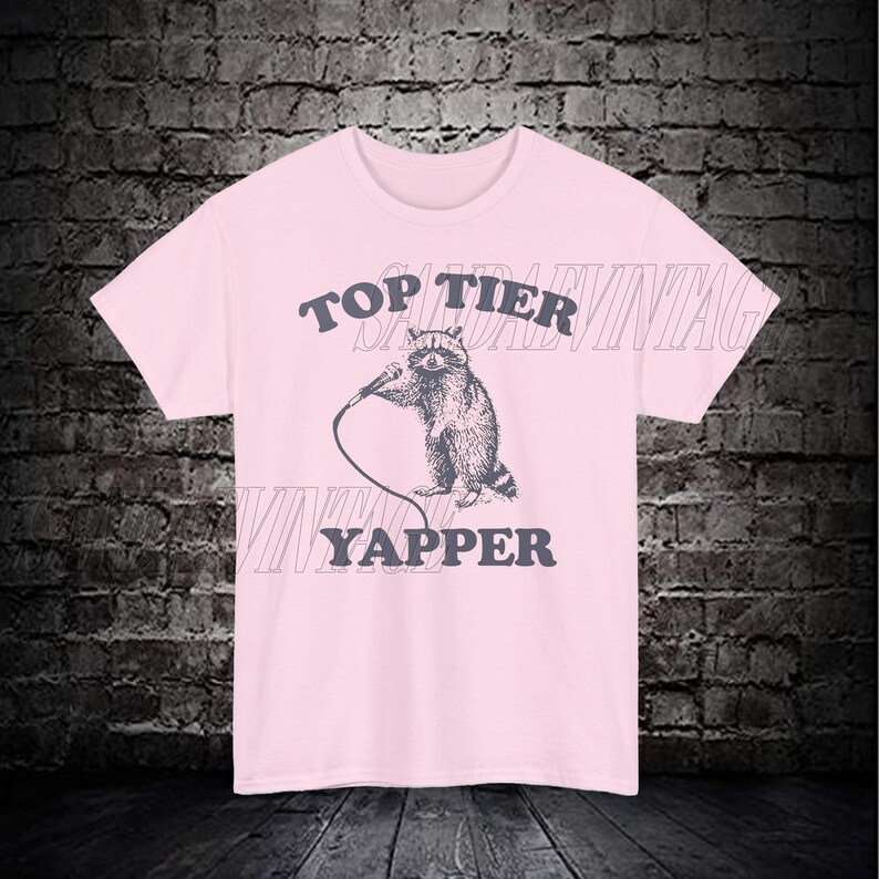May include: Pink t-shirt with a graphic of a raccoon holding a microphone and the text "Top Tier Yapper".