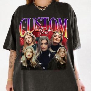 Peut inclure: T-shirt noir avec un motif graphique. Le motif présente le mot "CUSTOM" dans une police dégradée, et plusieurs images d'une personne aux longs cheveux blonds, sur fond rouge avec des éclairs.