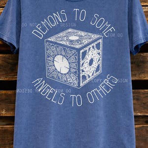 May include: Blue denim t-shirt with a white graphic of a cube and the text "DEMONS TO SOME, ANGELS TO OTHERS." The cube design has intricate patterns. The shirt is hanging on a wooden background.