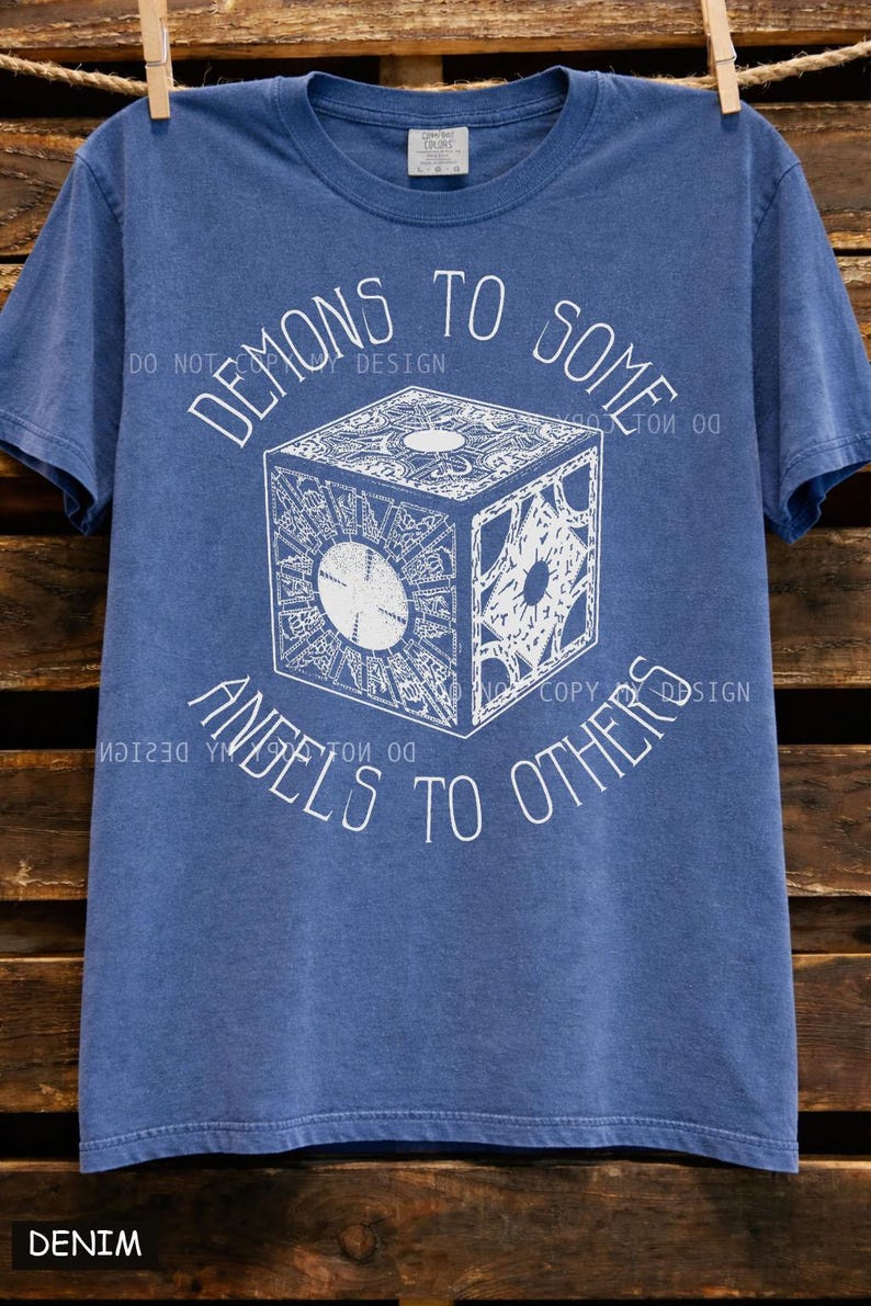 May include: Blue denim t-shirt with a white graphic of a cube and the text "DEMONS TO SOME, ANGELS TO OTHERS." The cube design has intricate patterns. The shirt is hanging on a wooden background.