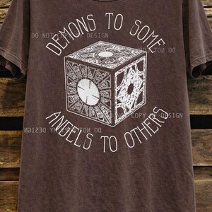 May include: Espresso-colored t-shirt featuring a detailed white graphic of a puzzle box with the text "DEMONS TO SOME, ANGELS TO OTHERS." The shirt is hanging on a wooden background.