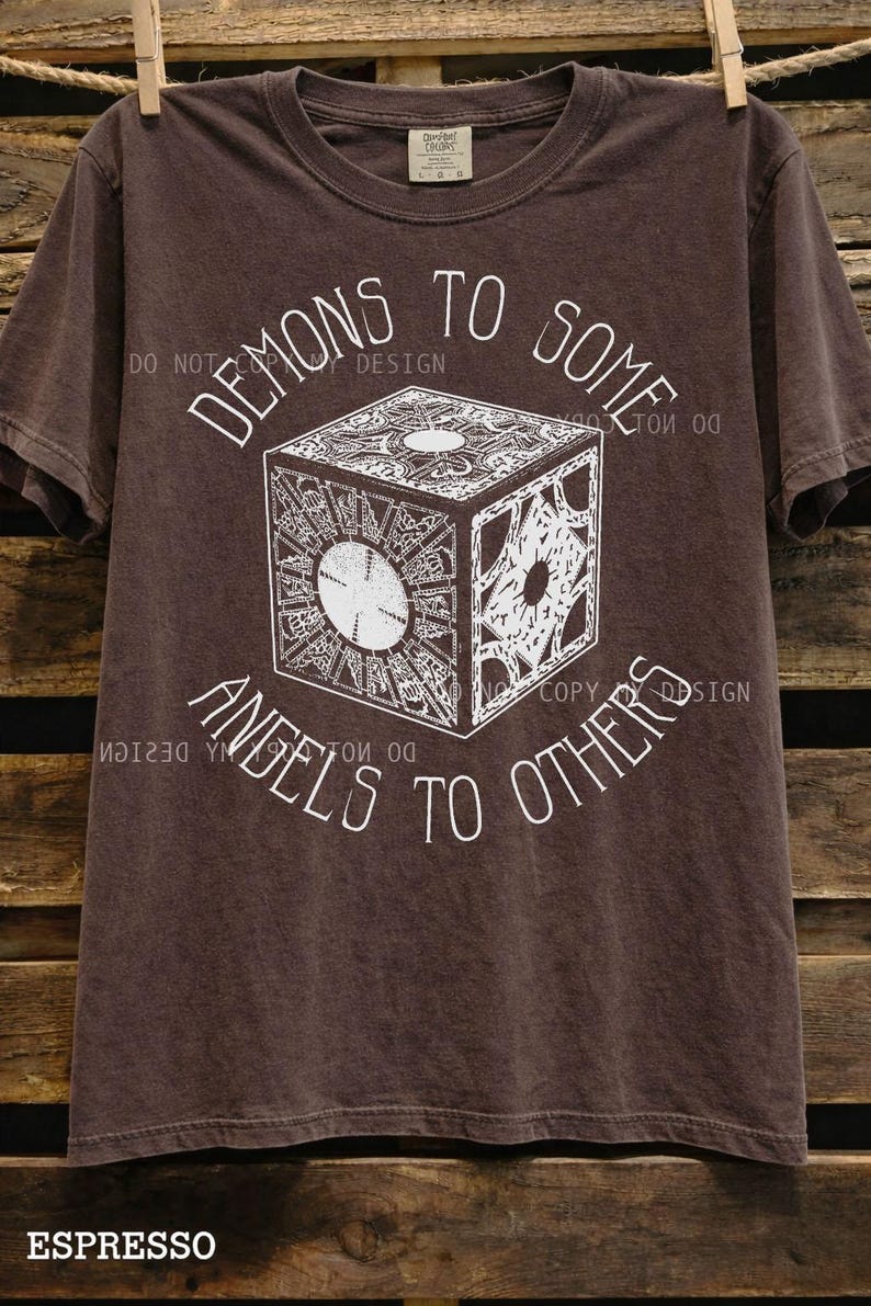 May include: Espresso-colored t-shirt featuring a detailed white graphic of a puzzle box with the text "DEMONS TO SOME, ANGELS TO OTHERS." The shirt is hanging on a wooden background.