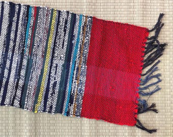 May include: A rectangular woven rug featuring wide solid red bands on both ends and a central section composed of multicolored vertical stripes in shades of navy blue, white, yellow, and teal, finished with dark blue fringe on the short edges.