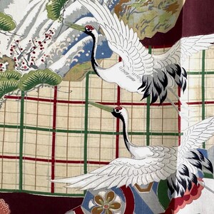 May include: A fabric with a Japanese-inspired design featuring two white cranes in flight. The cranes have black and gray wing details and red markings on their heads. The background includes a grid pattern in red and green, with landscape elements.