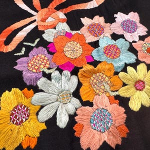 May include: A black fabric surface featuring a vibrant floral embroidery design. The pattern includes various multi-colored flowers in shades of orange, yellow, light blue, pink, and purple, accented by a long, flowing orange embroidered ribbon.