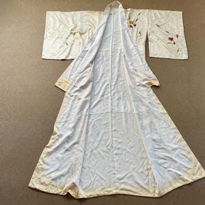 May include: Cream-colored kimono with floral embroidery in pink, yellow, and green. The long, flowing robe is laid out, showcasing its full length and wide sleeves. The fabric appears to be silk or a similar delicate material.