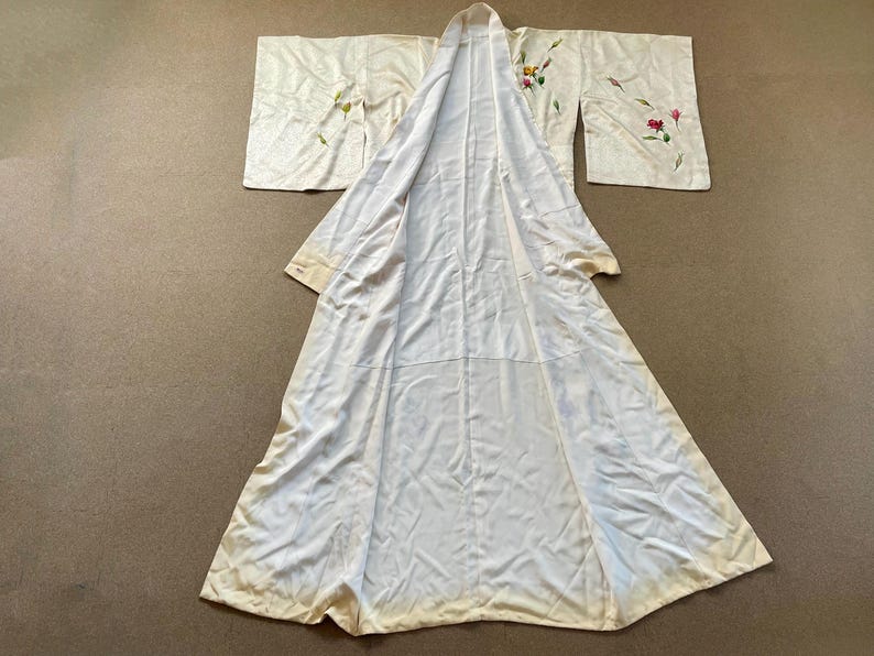 May include: Cream-colored kimono with floral embroidery in pink, yellow, and green. The long, flowing robe is laid out, showcasing its full length and wide sleeves. The fabric appears to be silk or a similar delicate material.
