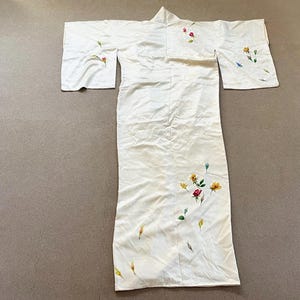 May include: Ivory-colored kimono with floral embroidery. The kimono features a long, flowing design with wide sleeves. Delicate embroidery of red, yellow, and blue flowers with green leaves adorns the garment, adding a touch of color and detail.