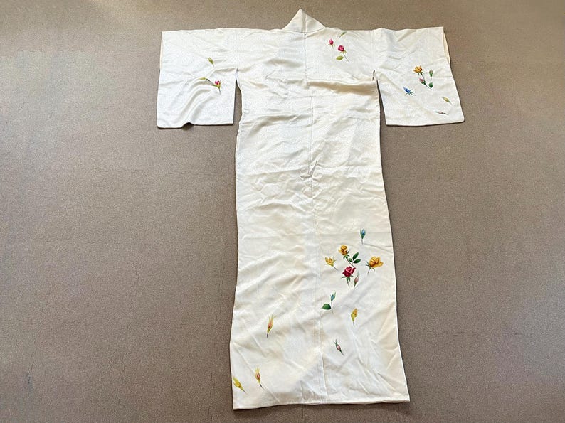 May include: Ivory-colored kimono with floral embroidery. The kimono features a long, flowing design with wide sleeves. Delicate embroidery of red, yellow, and blue flowers with green leaves adorns the garment, adding a touch of color and detail.