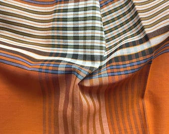 May include: Close-up of a folded fabric with a mix of patterns and colors. The fabric features orange, brown, and white stripes, along with a plaid pattern in green, black, and white. The fabric is a vibrant textile.