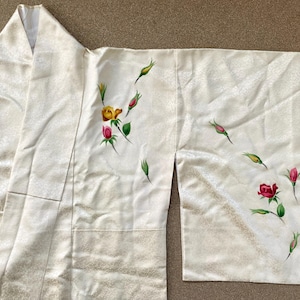 May include: Ivory-colored kimono with a floral design. The kimono features a pattern of roses in shades of red, yellow, and pink, along with green leaves and buds. The fabric has a subtle, textured appearance.