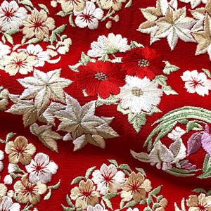 以下が含まれることがあります： 鮮やかな赤色の生地に、白、クリーム色、赤色の花の刺繍と、淡い緑色や金色のカエデの葉のモチーフが施されています。デザインには、紫と白の花びらが緑の葉に囲まれた円形のエンブレムが含まれています。