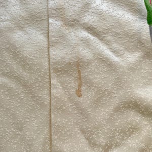 May include: Close-up of a cream-colored fabric with a subtle, textured pattern. A vertical seam runs down the center. A brown stain is visible, along with a partial image of a green and red floral design. The fabric appears to be a textile.