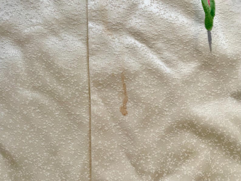 May include: Close-up of a cream-colored fabric with a subtle, textured pattern. A vertical seam runs down the center. A brown stain is visible, along with a partial image of a green and red floral design. The fabric appears to be a textile.