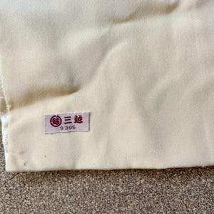May include: Close-up of a section of fabric in cream and white tones. A small rectangular label with Japanese text and the number 9395 is attached to the cream-colored fabric. The fabric appears to be part of a garment.