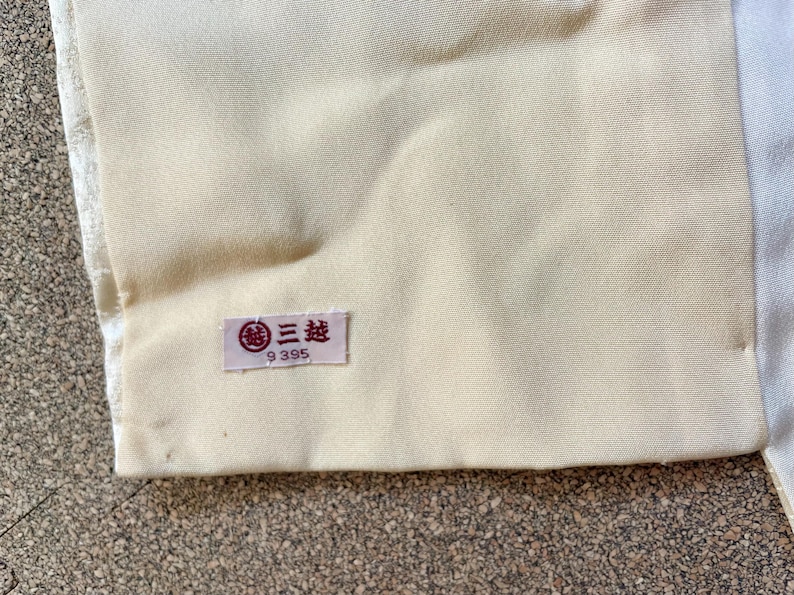 May include: Close-up of a section of fabric in cream and white tones. A small rectangular label with Japanese text and the number 9395 is attached to the cream-colored fabric. The fabric appears to be part of a garment.