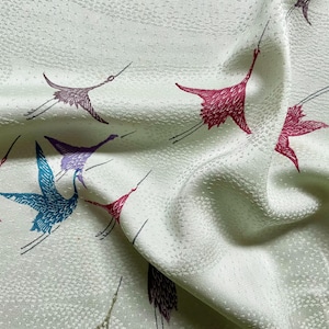 May include: Close-up of a light green fabric with a repeating pattern of flying cranes in shades of burgundy, blue, and purple. The fabric has a subtle textured surface, and the cranes are depicted in a stylized, minimalist design.