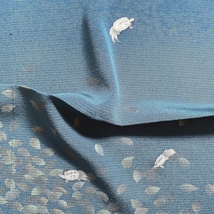 May include: Teal fabric with a pattern of white turtles and leaf shapes. The fabric has a textured appearance and is folded, creating shadows and depth. The design is repeated across the material.