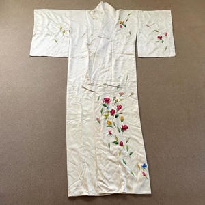 May include: Ivory-colored kimono with floral embroidery. The kimono features a long, flowing design with wide sleeves. Red, pink, yellow, and blue flowers are hand-painted on the front and sleeves. The garment is laid flat on a neutral surface.