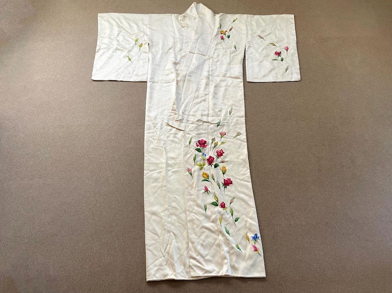 May include: Ivory-colored kimono with floral embroidery. The kimono features a long, flowing design with wide sleeves. Red, pink, yellow, and blue flowers are hand-painted on the front and sleeves. The garment is laid flat on a neutral surface.