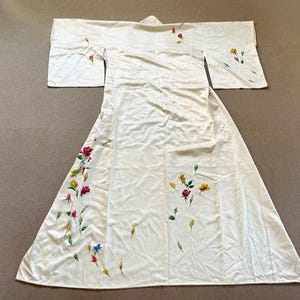 May include: Ivory-colored kimono-style robe with floral embroidery in red, yellow, and blue. The robe features wide sleeves and a floor-length skirt. The floral design is scattered across the robe's front and sleeves.