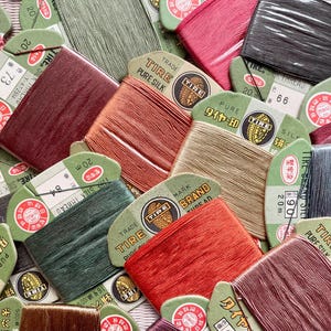 May include: A selection of vintage silk thread spools in shades of red, green, brown, and gold. Each spool features a paper label with Japanese text and the "TIRE" brand logo. The tightly wound spools are ready for use in craft projects.