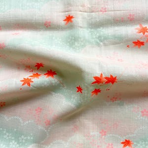 Può includere: Primo piano di un tessuto con sfondo verde chiaro e bianco. Il tessuto è decorato con motivi di foglie d'acero arancioni e fiori rosa. Il tessuto è stropicciato, mostrando la sua consistenza.