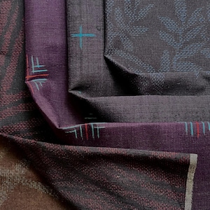 Op de afbeelding: Close-up van gevouwen stof in tinten paars, bruin en zwart. De stof heeft ingewikkelde patronen, waaronder geometrische vormen en bladachtige ontwerpen. Sommige secties hebben accenten van rood, blauw en wit.