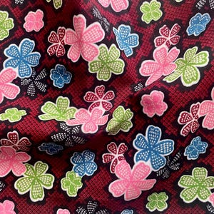 Puede incluir: Primer plano de una tela con un estampado floral repetido. Las flores son rosas, azules y verdes, sobre un fondo rojo oscuro con un diseño texturizado en forma de rejilla. La tela parece suave y fluida.