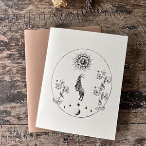 Könnte beinhalten: Eine Grußkarte mit einer Schwarz-Weiß-Illustration einer Ziege, Sonne, Sternen und Blumen in einem Kreis. Dazu ein brauner Umschlag, auf einem rustikalen Holzgrund.