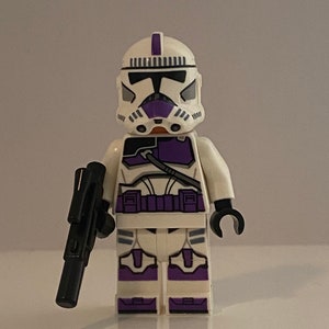 May include: A white Lego minifigure of a clone trooper with purple accents. The figure has a black blaster rifle in its left hand.