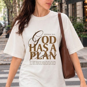 Puede incluir: Una camiseta blanca de manga corta con texto marrón que dice God Has A Plan Jeremiah 29:11, junto con el texto completo del versículo debajo. La camiseta se lleva con un bolso de hombro marrón, mostrando un diseño sencillo y casual.