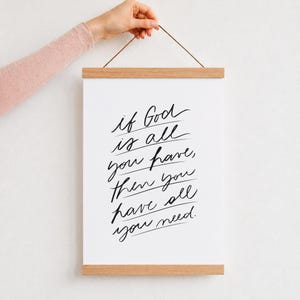 Puede incluir: Un cartel rectangular blanco con texto en cursiva negra que dice: if God is all you have, then you have all you need. La impresión está asegurada entre dos listones magnéticos de madera clara y suspendida por un cordón marrón fino.