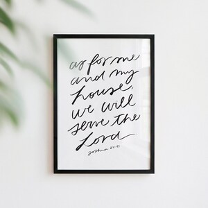 Puede incluir: Un cuadro decorativo con marco negro que presenta un fondo blanco y caligrafía manuscrita en negro con el texto: as for me and my house, we will serve the Lord. Joshua 24:15. Las letras están escritas en un estilo cursivo fluido.