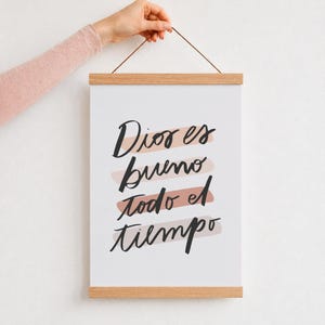 Puede incluir: Un cartel vertical para colgar en la pared con un fondo claro y el texto manuscrito en negro 'Dios es bueno todo el tiempo' sobre trazos de pincel rosa suave. La lámina está sujeta entre dos listones magnéticos de madera clara.