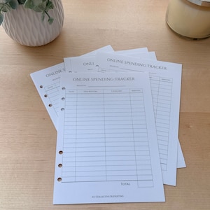 May include: Several white paper sheets with the text "ONLINE SPENDING TRACKER" are stacked on a light wood surface. Each sheet has a grid for tracking expenses, with columns for date, description, category, and amount. A white plant pot and a candle are in the background.