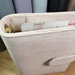 Może przedstawiać: Jasnor&oacute;żowy, teksturowany sk&oacute;rzany planer ze złotym zapięciem i pierścieniami. Wewnątrz widoczna jest karta z napisem "DAILY". Tło przedstawia inne planery w r&oacute;żnych pastelowych kolorach.
