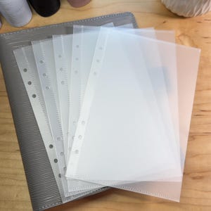 May include: A stack of clear, translucent plastic binder sleeves with punched holes, fanned out over a gray, textured binder. The sleeves are designed to hold papers and documents. The background is a light-colored wooden surface.