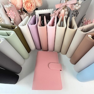 May include: A collection of textured, faux leather personal planners in various colors including pink, white, lavender, olive green, mint, beige, gray, and black. Each planner features a secure snap-button closure and a structured, upright design.