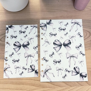 May include: Two planner inserts with a repeating pattern of gray bows on a white background. The inserts have pre-punched holes for binding. The bows vary in size and are shaded to give a 3D effect.
