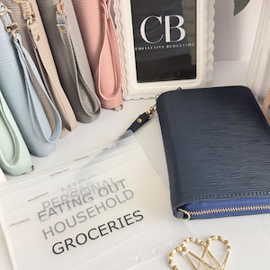 May include: A collection of pastel-colored wristlet wallets with gold zippers and wrist straps. A navy blue wallet, clear budget dividers, and gold paper clips are also present. The dividers are labeled with budget categories like "Groceries" and "Household."