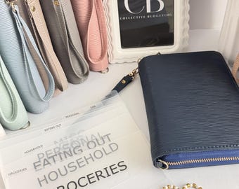 May include: A collection of textured zip-around wallets in light blue, gray, pink, and navy blue, each with a wrist strap. Included are translucent plastic cash envelopes labeled Groceries, Household, Eating Out, and Personal, plus gold paperclips.